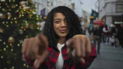 Woman in red plaid shirt points finger at camera on busy city street by christmas tree; friendly...
