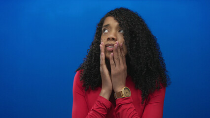 African american woman with tightly clasped hands near lips in blue studio, eyes closed in worried...