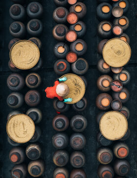 Aerial view of a person with a red conical hat amidst rows of earthenware pots, creating a pattern of rustic browns and earthy tones, Soy sauce maker, Ban Yen Nhan village, Hanoi Vietnam.