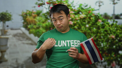 Young man holding thailand flag in green volunteer shirt outdoors with urban backdrop and potted...