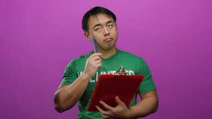 Young man standing with clipboard wearing green shirt against pink wall isolated background thinking thoughtful.