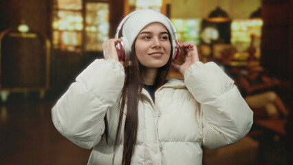 Woman enjoying music in cozy hotel lobby wearing headphones and winter clothing, capturing a moment...