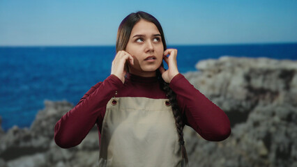Hispanic waitress wearing an apron covers her ears by the seaside with a serene beach and sea...