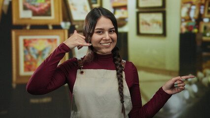Young waitress in apron at art gallery gestures pointing and mimics phone call surrounded by vibrant paintings.