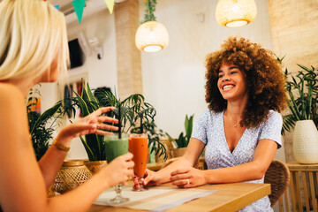 Two happy female friends are drinking smoothies at a bar and chatting together