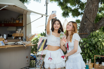 Two women are standing in front of a food truck. Happy asian LGBTQ lesbian couple on a summer vacation, taking selfies and relaxing at a tropical beach cafe.