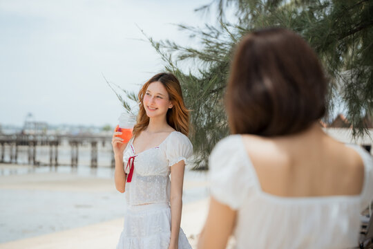A woman is standing on the beach with a drink in her hand. Two happy diverse women friends taking a selfie on the beach during a summer travel vacation.
