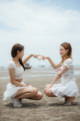 Two women are sitting on the beach and making a heart shape with their hands. One of the women is wearing a white dress