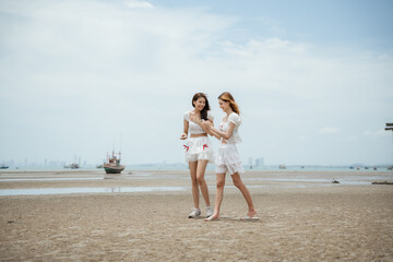 Two women are walking on the beach. One of them is holding a cell phone. The other woman is holding a handbag