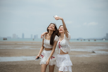 Two women are standing on a beach, one of them taking a picture of the other. They are both wearing white clothing