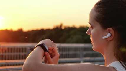 A young woman wearing a white tank top, earbuds, and a fitness tracker checks her wrist, with the setting sun and a blurred background. - Powered by Adobe