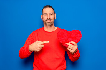 Bearded Hispanic man in his 40s wearing a red sweater holding a red heart as a symbol of romance and health, looking surprised and pointing at himself with his finger. Isolated on a blue background.