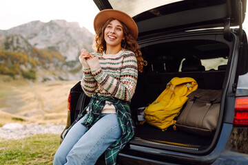 A cute woman on a road trip sits in the trunk of a car and enjoys the mountain scenery. A female...