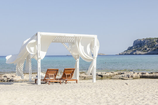 View of sun-drenched beach cabana with billowing white curtains frames a serene turquoise sea, inviting relaxation and escape, Lindos, Rhodes, Greece.