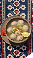 Bakso served in a traditional clay bowl with chili sauce and lime on the side, on a batik tablecloth, top-down view, food styling photography