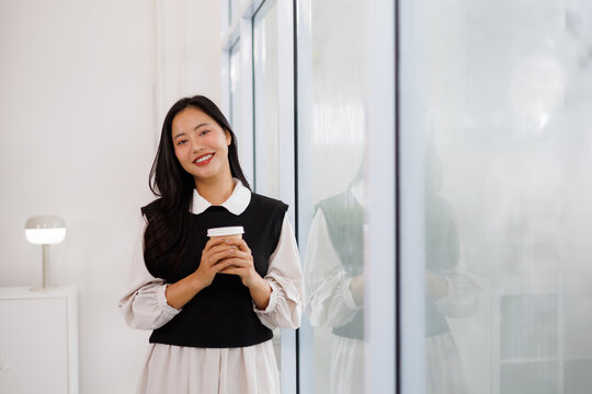 Young asian businesswoman smiling and holding coffee cup in modern office - Powered by Adobe