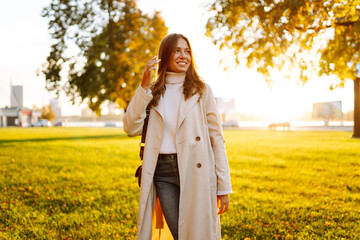 A young woman stands in a sunny park and enjoys a walk. A beautiful woman in stylish clothes relaxes outdoors. Walking, beauty and fashion concept.
