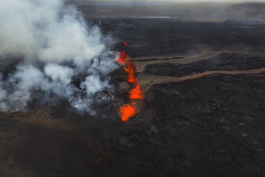 Aerial view of molten lava snaking through the dark, volcanic landscape, a fiery river contrasting with the cool, ethereal smoke, Grindavik, Southern Peninsula Region, Iceland.