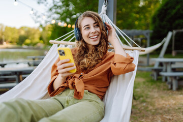 Smiling woman with headphones and phone lying in hammock enjoying weekend in sunny park. Beautiful woman in hammock listening to music or podcast outdoors. Technology concept, weekend.
