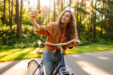 Cheerful woman with phone and bicycle taking selfie in sunny park. Young woman riding bicycle and enjoying weather outdoors. Walking concept, selfie. Active lifestyle.