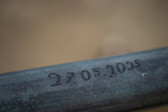 Macro shot of a metal bar with the date "27.05.2025" engraved on it, set against a softly blurred brown background.