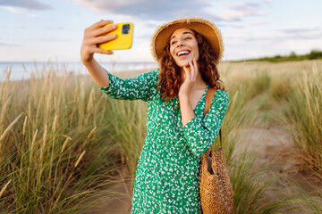 Cheerful woman in a hat takes a selfie on phone against backdrop of a sandy beach. A beautiful...