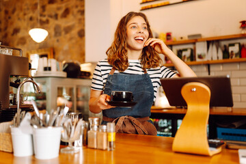 A young female barista stands behind a bar counter with prepared coffee. A beautiful woman with a...