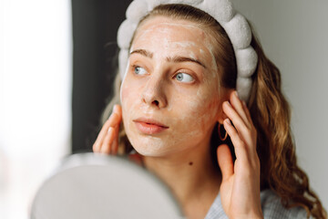 A young woman takes care of her skin by applying a face mask and looking at a small mirror in a...