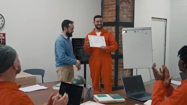 Zoom-out shot of proud Middle Eastern male convict in orange uniform receiving certificate for successfully completing computer programming course during rehabilitation while serving jail sentence