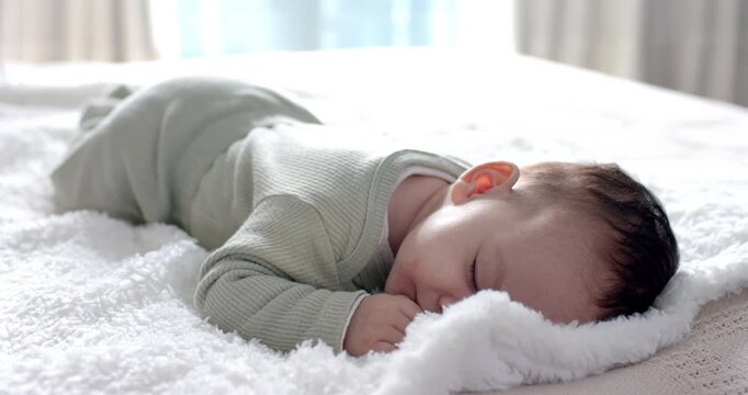 Infant female baby wearing pale green onesie napping on white blanket in bedroom breathing gently