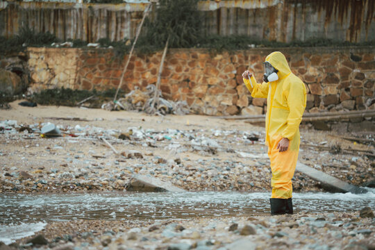 Water quality inspection, Environment scientist in hazmat suit holding a water sample vial in a polluted industrial area. Concept of environmental analysis, factory wastewater, and hazard assessment