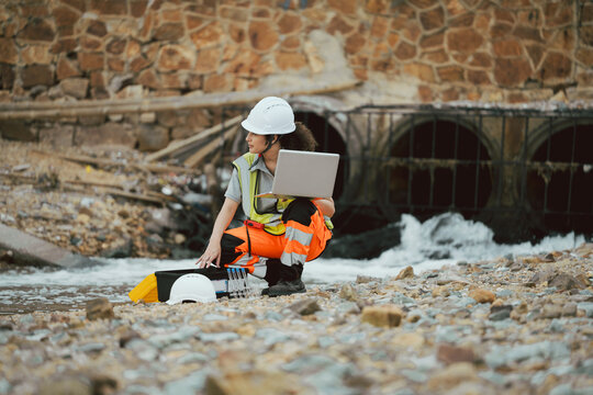 Female engineer with laptop inspecting water drainage pipes at an industrial site. Concept of environmental monitoring, infrastructure management, and technology in engineering
