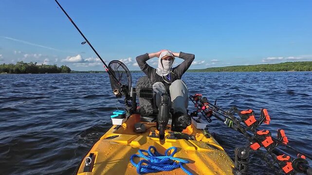 Fisherman in waders and sunscreen hoodie relaxing in solitude after bass fishing competition. Angler pedaling motorizing kayak to the boat launch at hot afternoon. Catching fish game tournament.