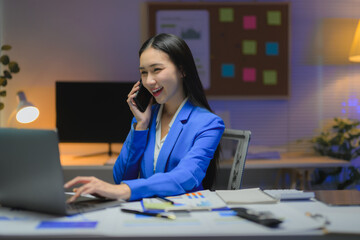 Smiling asian businesswoman talking on phone and working on laptop at night