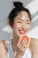 A natural-looking young Korean woman with glowing skin, softly tied messy bun, wearing a white sleeveless top, smiling while holding a slice of grapefruit close to her mouth.