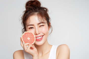 A natural-looking young Korean woman with glowing skin, softly tied messy bun, wearing a white sleeveless top, smiling while holding a slice of grapefruit close to her mouth.