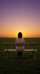 Woman sitting alone on a park bench watching the sunset. Solitary female figure in a vast, empty field. Contemplation and loneliness concept.