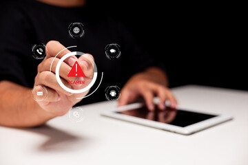 A hand points to a digital warning symbol for a scam, with an electronic tablet on a white table in the background.
