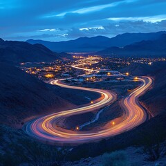 A winding mountain road illuminated by streaks of car lights at dusk, with a city glowing in the valley and mountains silhouetted against a blue sky.