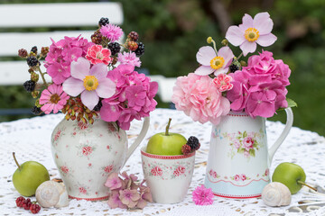 florales Arrangement mit Blumensträußen mit pink Hortensienblüten, Herbst-Anemonen, Rosen, Nelken und Zinnie in Porzellan-Kännchen