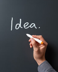 A hand writes the word "Idea." in white marker on a dark chalkboard or blackboard surface.