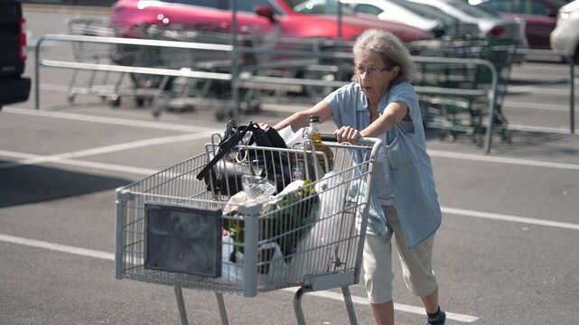 An elderly woman is shopping at a grocery store, pushing her cart filled with food items across the empty parking lot on a sunny day.