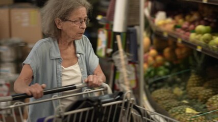 An elderly senior woman browses produce in a grocery store, examining avocados amid a colorful display of fruits. She is focused on her selections. - Powered by Adobe