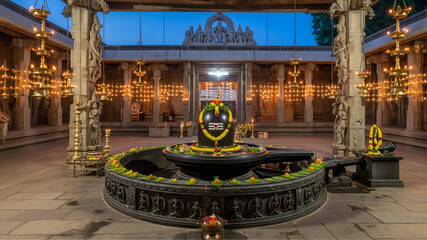 Large black stone Shiva Linga in a South Indian temple courtyard with hanging oil lamps and stone pillars