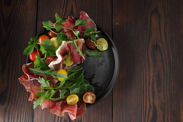 Black plate with thinly sliced jamon, colorful vegetables and greens.