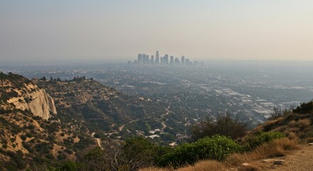 Obraz premium Distant cityscape view of Los Angeles skyline shrouded in haze, seen from a hilly landscape with vegetation.