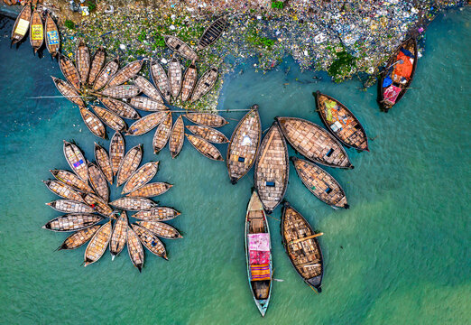 Aerial view of boats clustered near a trash-filled shoreline, the turquoise water contrasting sharply with the colorful vessels, Dhaka, Dhaka District, Bangladesh.