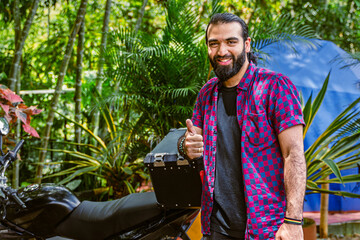Person standing next to motorcycle in tropical setting, looking at camera with thumbs-up gesture,...