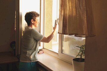 Smiling woman takes a photo of the city skyline with her smartphone from an open window in warm evening light, enjoying the sunset and peaceful home atmosphere