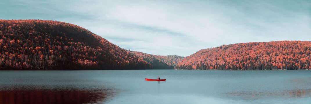 Man in red canoe on calm lake with stunning autumn forest in background. Vintage outdoor adventure and travel concept for fall season. Vintage style, fine grain.